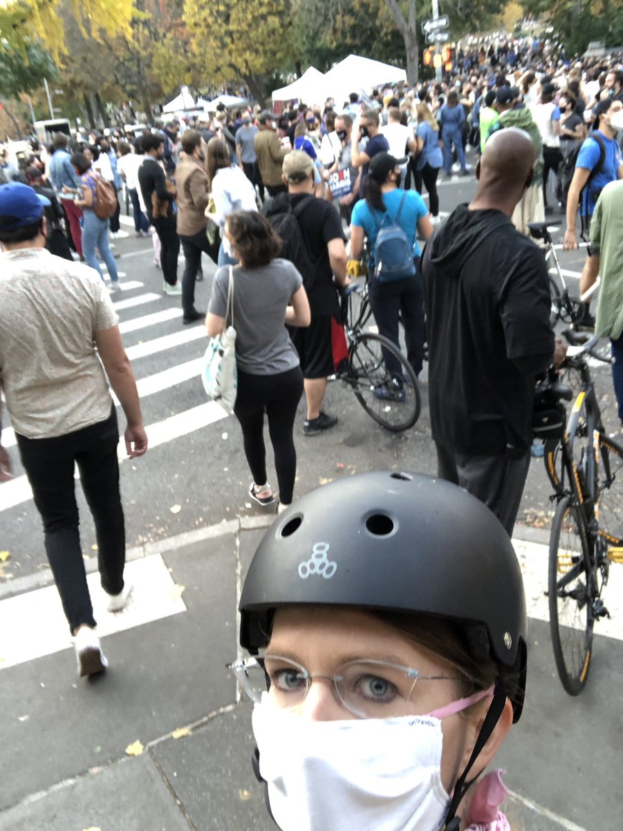 Chelsea E. Manning stands on the corner of a street and busy road crossing with a busy park of excited people while wearing a bicycle helmet and cloth mask in Brooklyn after celebrating Donald J. Trump losing the election, and failing to gain another term of the U.S. Presidency