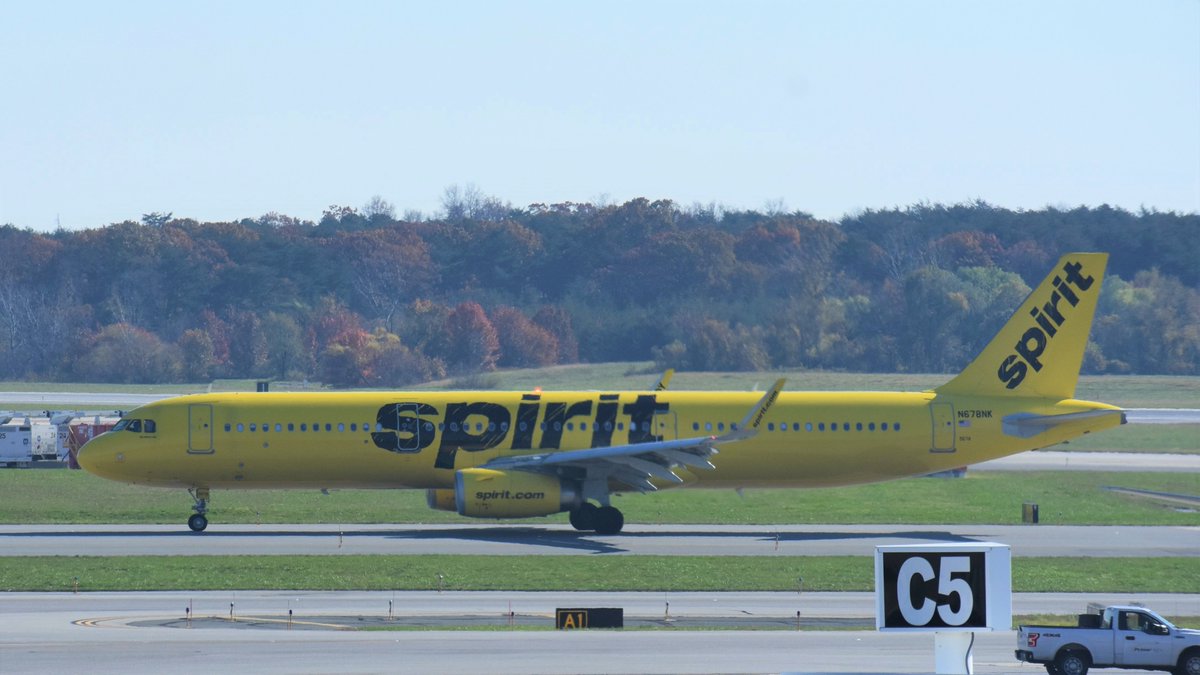 Photograph of Spirit Airlines aircraft on taxiway in front of trees with fall color foliage in background.