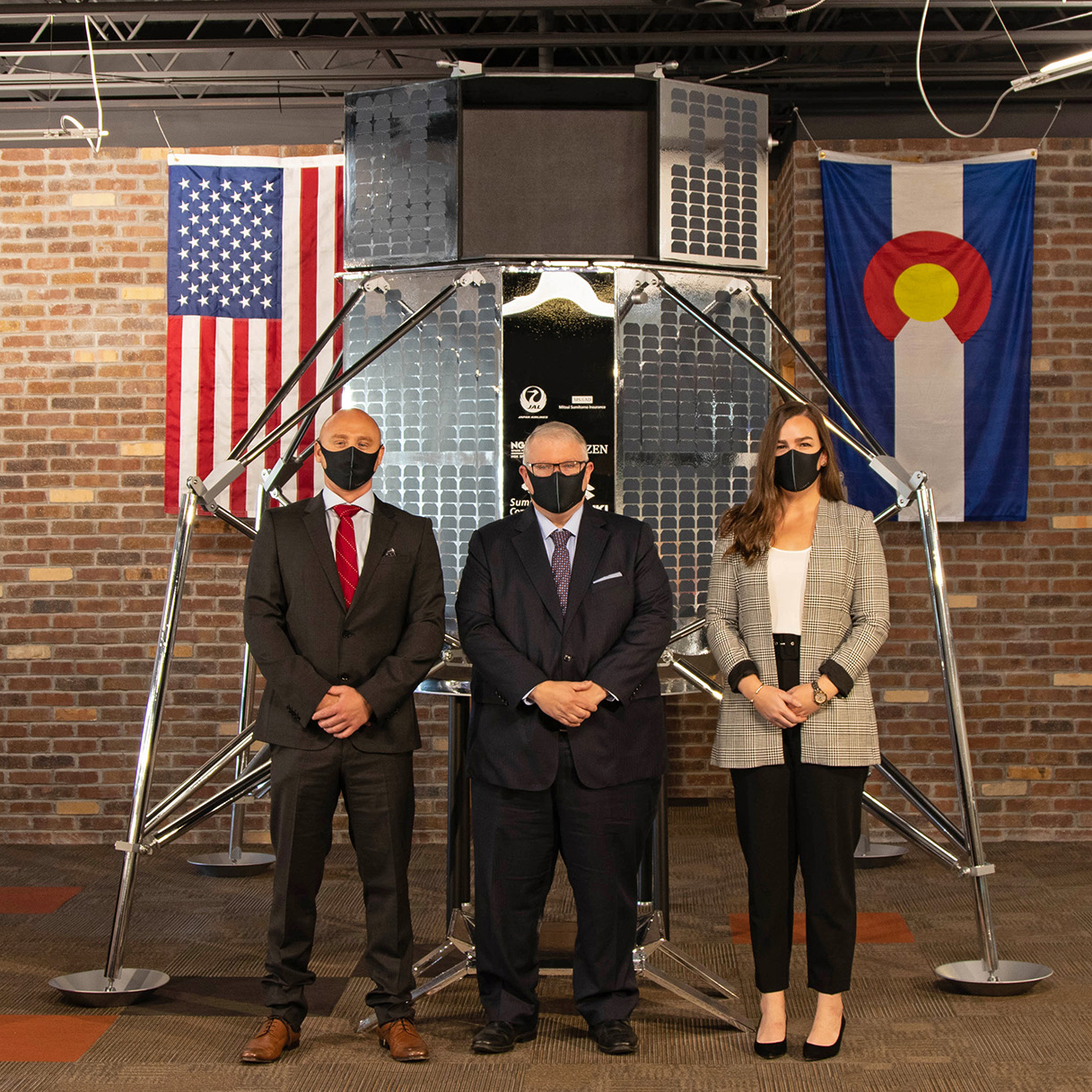 Kevin O'Connell and two others standing in front of a lunar lander with U.S. and Colorado flags in the background