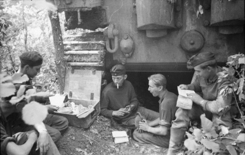 Heer Tiger crew in typical laager setting, with sleeping pit/shelter under the vehicle. Note the recycled box with the crew’s all important mess kit & eating utensils as the men read letters from home? The smoker wears a civilian jumper. Date estimate 42/43. 43)