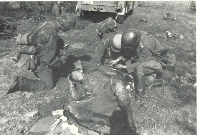 An Assistenzarzt & Sanitäter treat a serious arm injury in an area seemingly behind the HKL. The Assistenzarzt has forceps as the Sanitäter provides support. The Dr’s medical Tornister pack lies at the casualty’s feet & the soldier to the left has water at the ready.48)