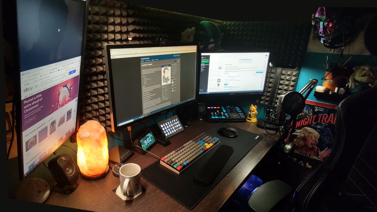 A glorious 4K monitor is centre, flanked by a vertical monitor to the left and standard horizontal to the right. A warming salt lamp, sits, embering away next to a colourful Vortex Tab 90M keyboard. Strewn around are foam wall panels, devices, a microphone and the faint colour of the PC glowing underneath the desk. This is a place of comfort, of productivity, of safety.