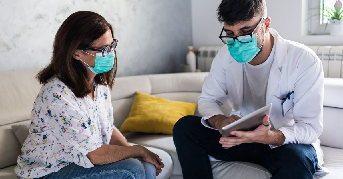 Male consultant sitting on white couch in home with female client - both wearing masks. Consultant is holding out tablet to demonstrate process to the client.