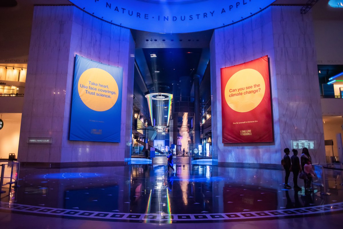 Image of the Museum of Science and Industry's large Rotunda, with its Science Storms exhibition in the background showing an indoor tornado. Banners flanking the exhibit read "Take heart. Use face coverings. Trust science," and "Can you see the climate change?"