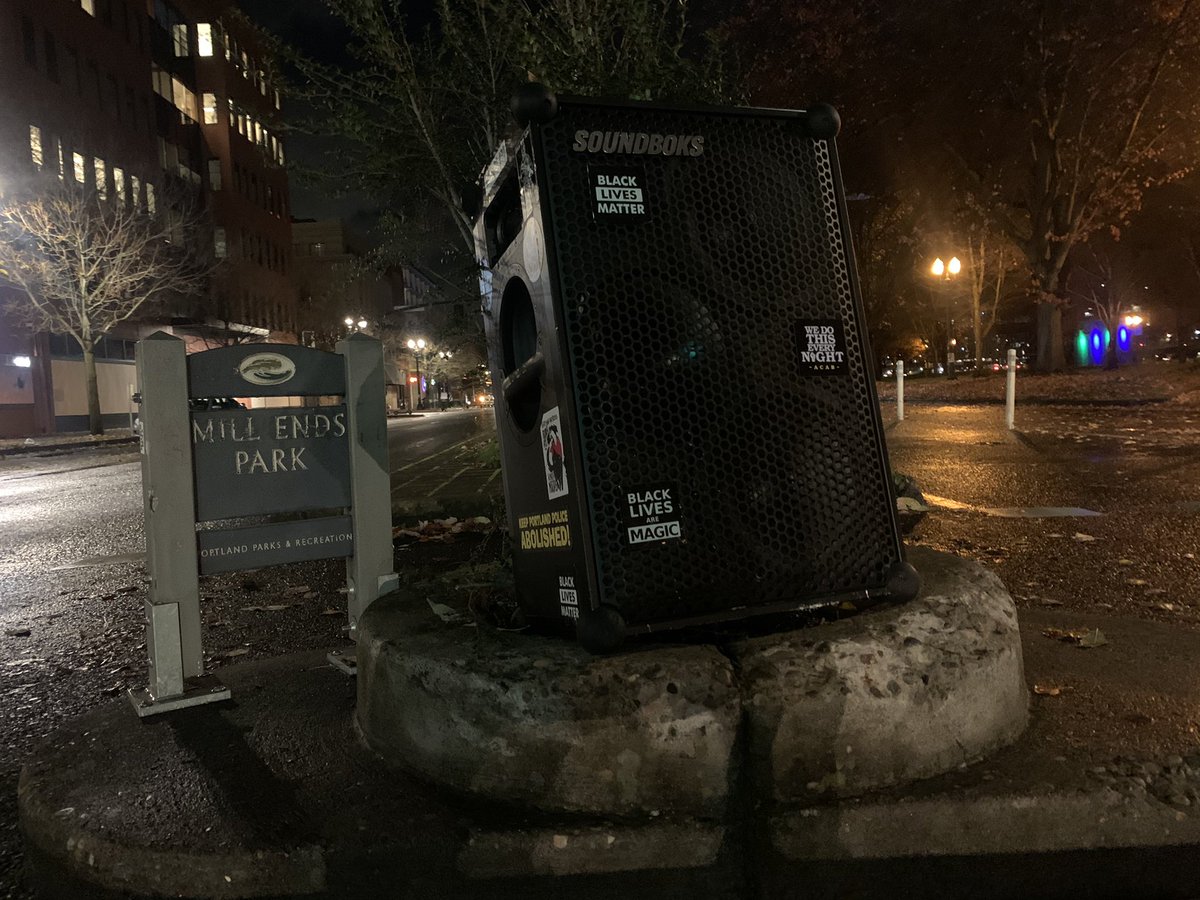 Mills end park sign on the left with the speaker on top of the scraggly bush in the center. Still in the middle median of street. The ground is wet and glistens with fall leaves scattered.