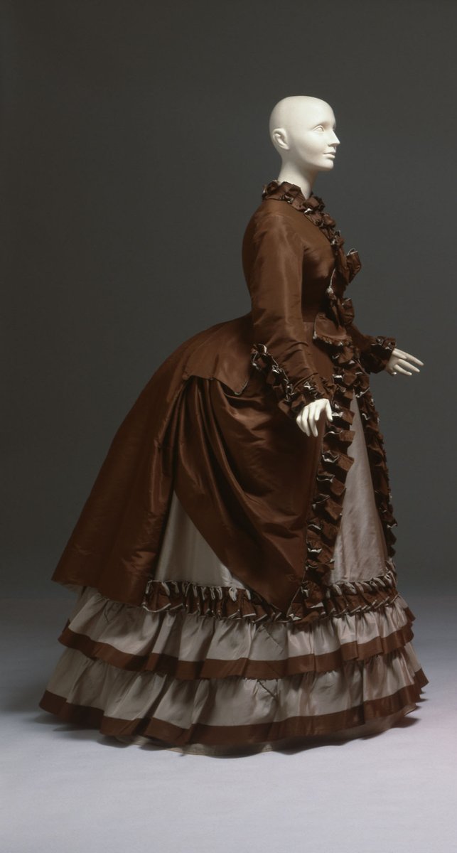 The transition from the voluminous crinoline-enhanced skirts of the 1850s and 1860s can be seen in the loops and gathers of fabric and trimmings worn during this period.