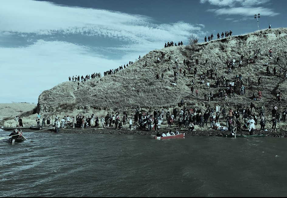 Photograph of a large group of people standing on a hill at the Standing Rock reservation. The hill overlooks a body of water. Many people are standing on the banks, and some are in canoes within the water itself. The sky is blue with white clouds in the background. 