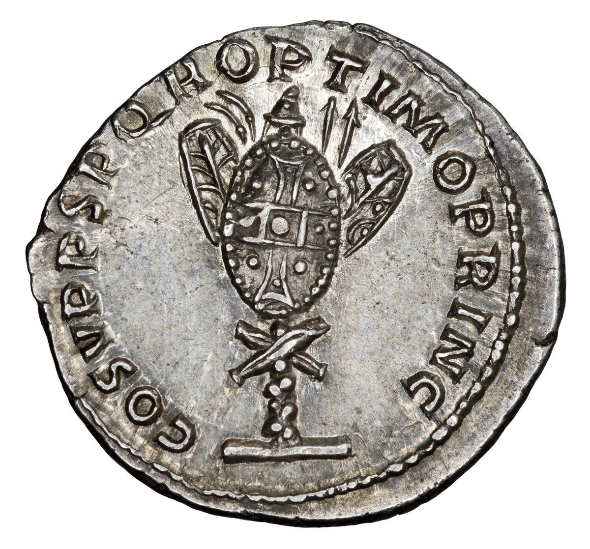 The trophy on this denarius is instead mainly constructed of highly decorated Dacian shields; one large, round shield at centre with hexagonal ones either side. Behind we see javelins and the distinctive curved Dacian falx. Atop is a helmet and below, a pair of crossed greaves.
