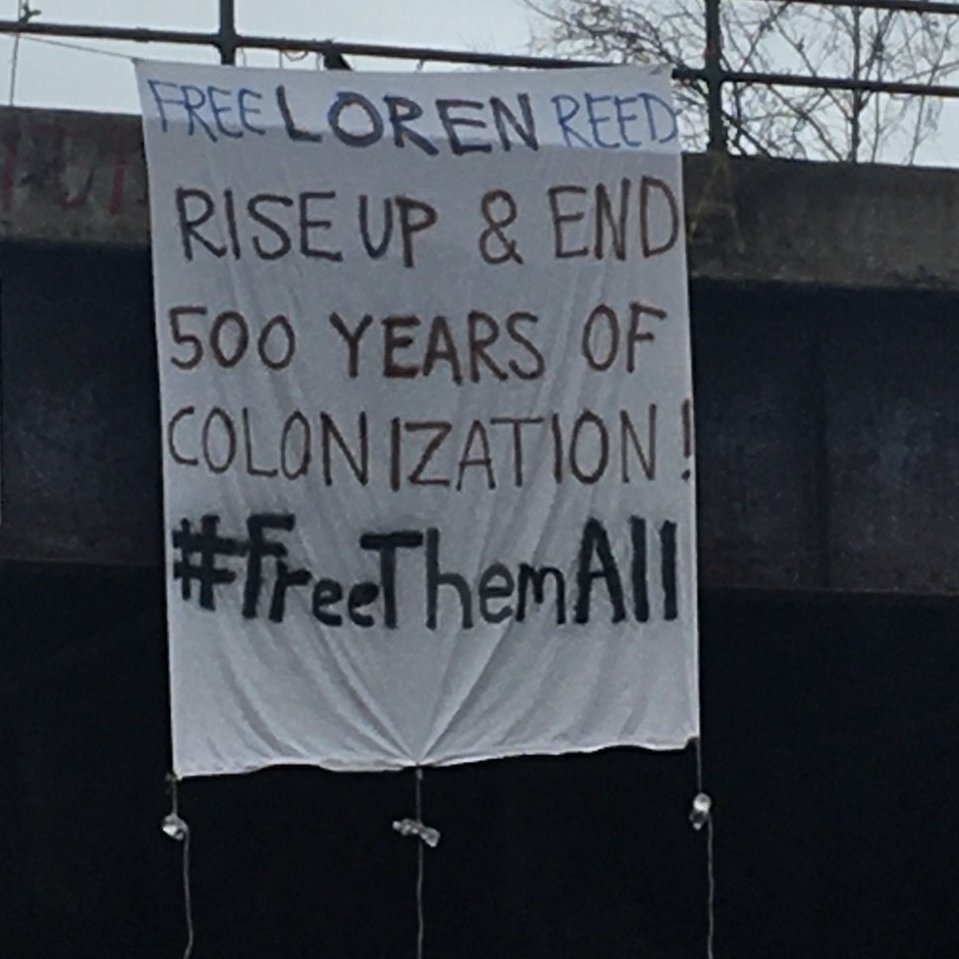 Banner reading "Free Loren Reed. Rise up and end 500 years of colonization" with a hashtag that reads "Free Them All."