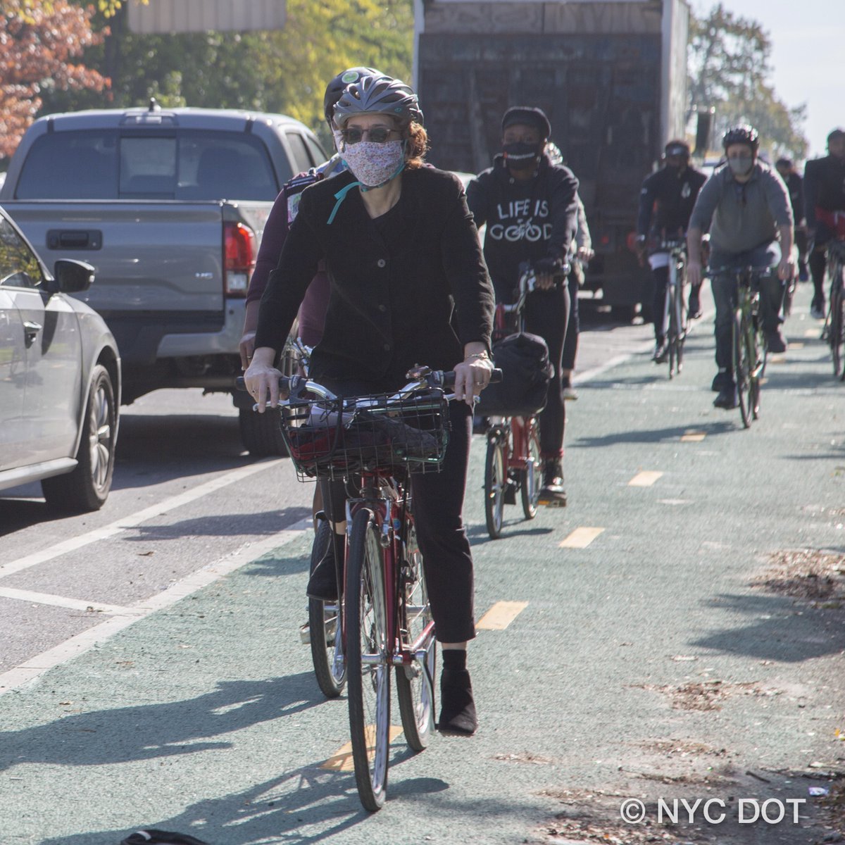 Commissioner Trottenberg, wearing a helmet and a mask, rides a bike in a green bike lane. A person behind her wears a shirt that says life is good.
