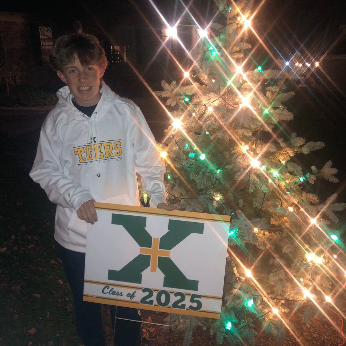 StX_Principal's tweet image. This made my day! Sam Shewmaker, Class of 2025, had his picture taken by his neighbor and St. X alum's green/gold tree. Thank you, Jim Head '50, for keeping those lights burning every year. Welcome home, Sam! #WeAreStX