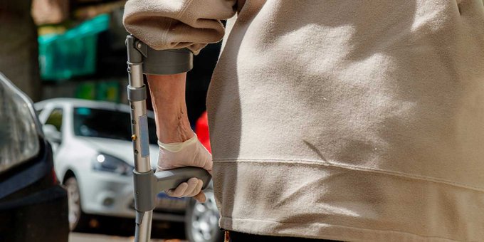 Cerca Fotografía en primer plano de la espalda y el brazo izquierdo de una persona que se prepara para cruzar una calle. La persona está utilizando una muleta