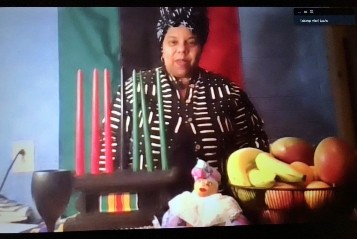 Micki Davis standing in front of a Kwanzaa kinara and bowl of fruit with a flag behind her