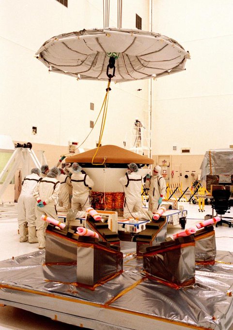 Photo of a roughly cylindrical piece of machinery raised on a small platform in a large warehouse. Several people in white coveralls are crouched on the platform working on the machine, and the top of the machine (a large disk) is suspended overhead.