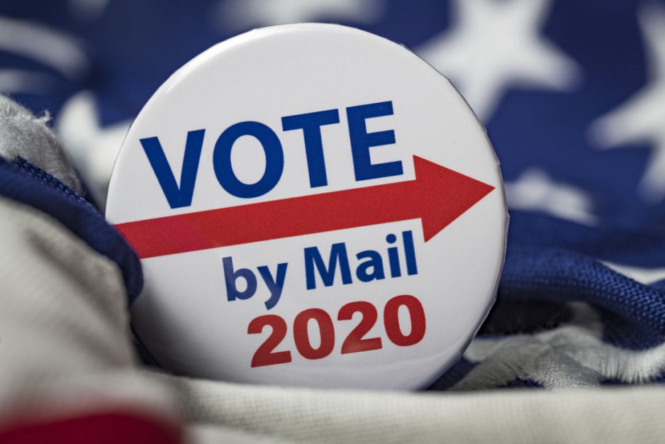 Photograph of round, white, pin-on button with the words "Vote by Mail 2020" printed in blue and red with a red arrow. 