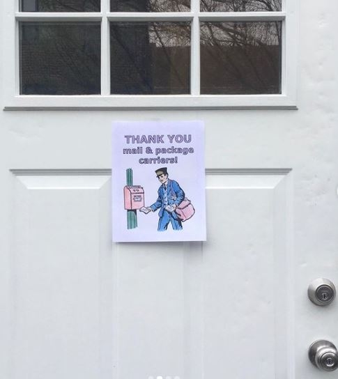 Image of a white front door with windows and a silver lock and knob. Taped to the door is a white piece of paper with a crayon drawing of a male letter carrier mailing a letter. Text reads "Thank you mail & package carriers."