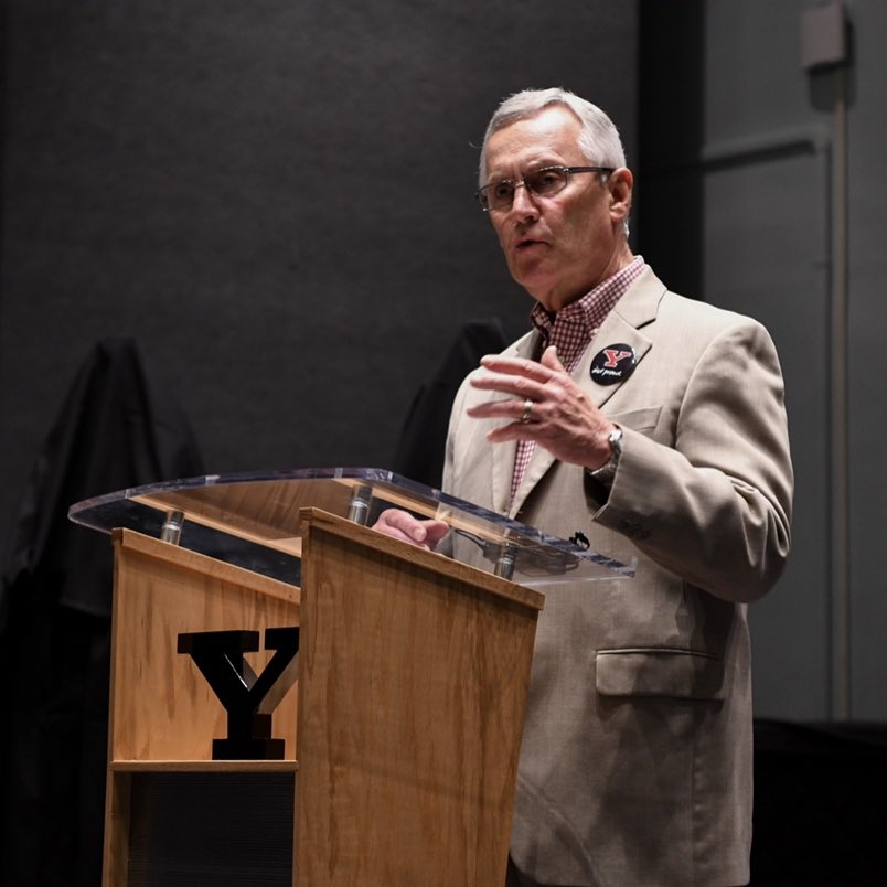 Jim Tressel stands at a podium labeled for Youngstown State University, speaking to a crowd not shown in view.