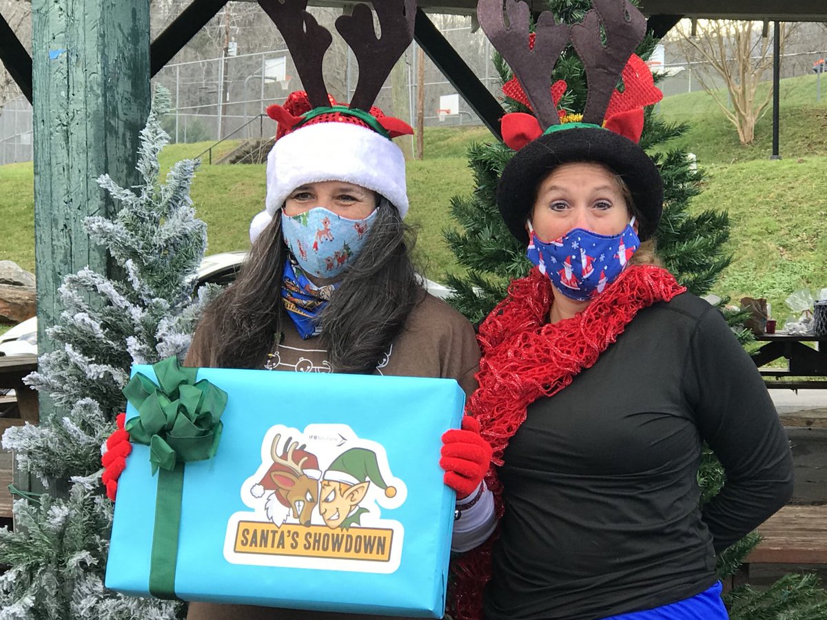 (Image: Two women dressed in reindeer antlers smile while wearing masks holding a gift box wrapped with a Santa's Showdown logo across the front).