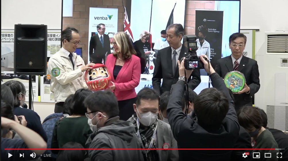 Yuichi Tsuda is holding a daruma; a Japanese luck doll. The eyes are initially blank. One is drawn in to represent a goal, while the 2nd is added once that goal is fulfilled. The control room has a line of daruma representing different operations, but I think this is the biggest.