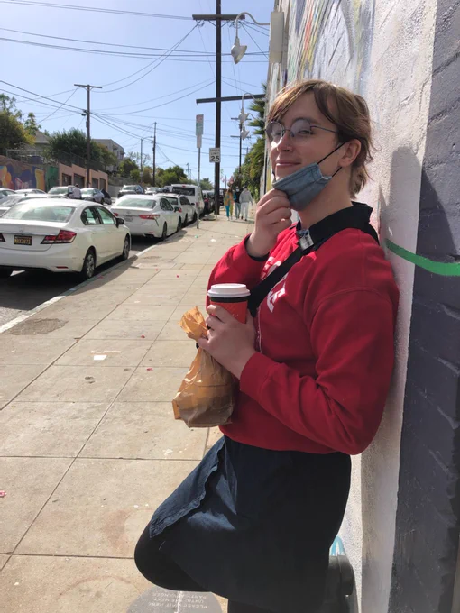 Me, leaning against a wall in a denim dress and black boots with a red crew neck, I'm looking at the camera with the mask pulled down just enough to see my smile
