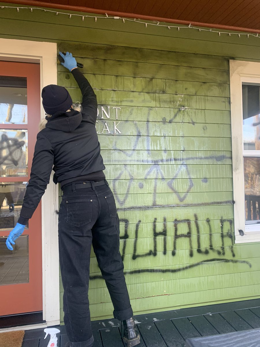 a person wearing black stands in front of a green wall with their back to the camera. they’re holding a brush and scrubbing black graffiti off the wall. the graffiti reads “Nazis go to Valhalla”