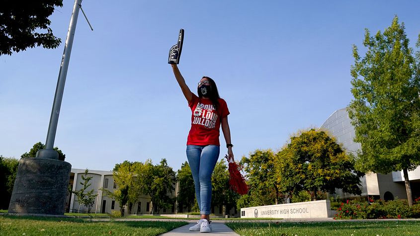 A Fresno State student sports a Fresno State spirit shirt while waving pompoms and a foam finger. 