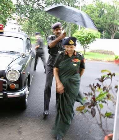 A uniformed trooper runs to the rear door with an open umbrella to shield the officer who steps out. Dressed in the Indian Army's olive green uniform, the officer pulls the pleats of her sari and walks up. +