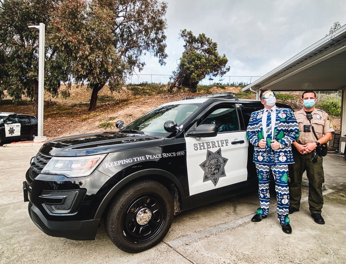 Detective Reed dressed in a Christmas themed suit posing his best next to a patrol car and Sgt. Houser.
