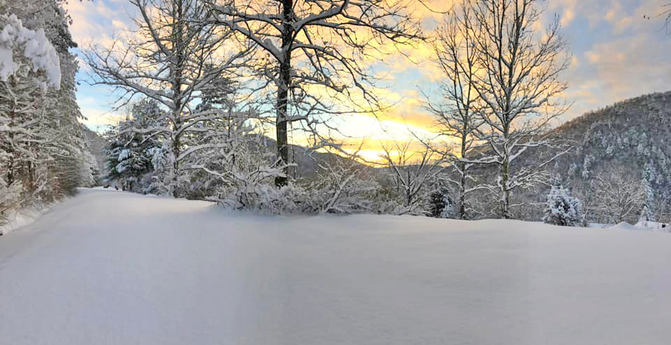 Sunrise brightening sky in distance state forest road, trees, and mountains covered in deep snow.