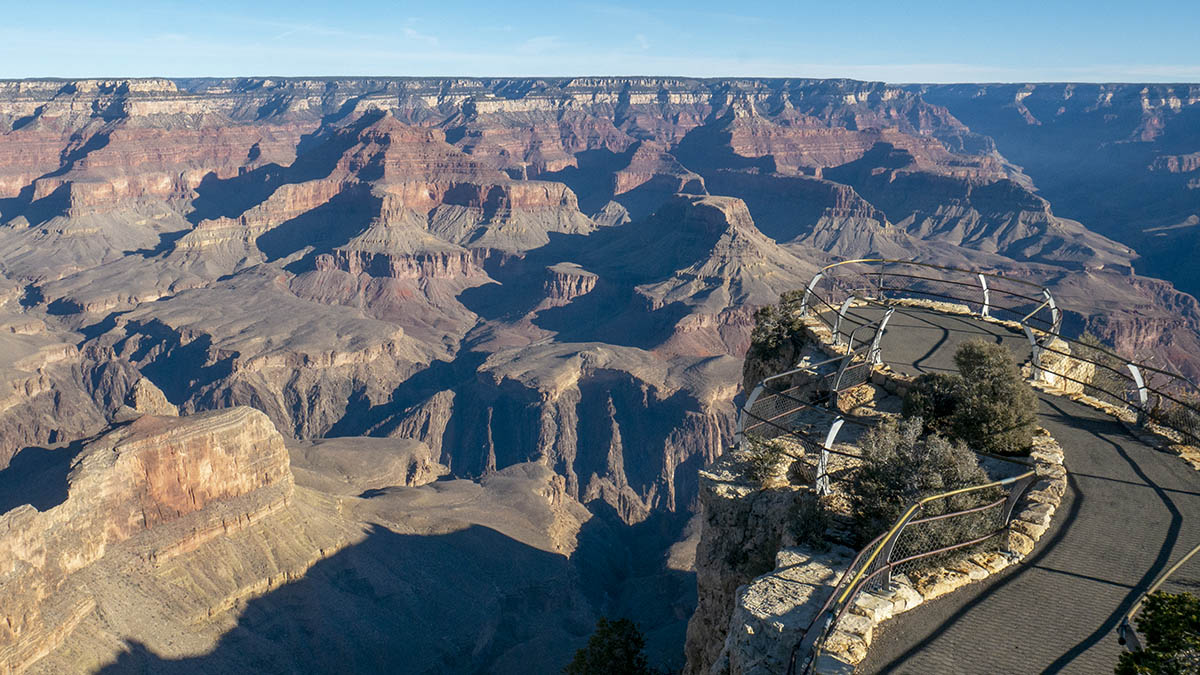 Shortly after sunrise, colorful peaks and cliffs are seen rising from the floor of a mile-deep canyon. A narrow scenic overlook in the foreground with guardrails on all sides.
