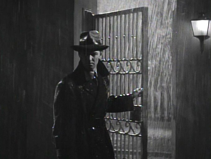 Alan Ladd dressed in overcoat and hat, opening a gate at night, in the pouring rain