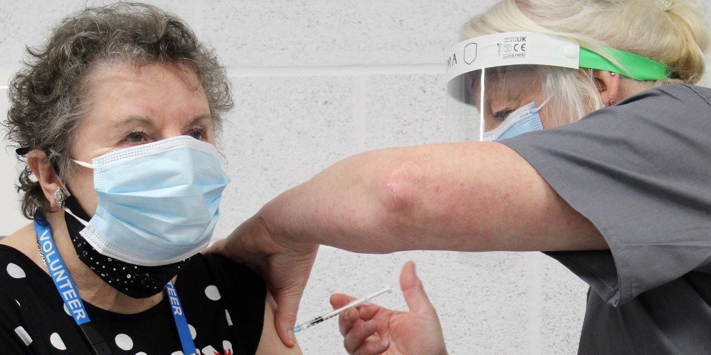 Norma is receiving her COVID-19 vaccine from a nurse wearing a visor and face mask. Norma is looking slightly off camera and has a blue medical face mask on. 