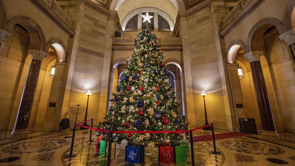 A large Christmas tree with lights and ornaments sits in the central rotunda at Los Angeles City Hall. A menorah is visible in the background.