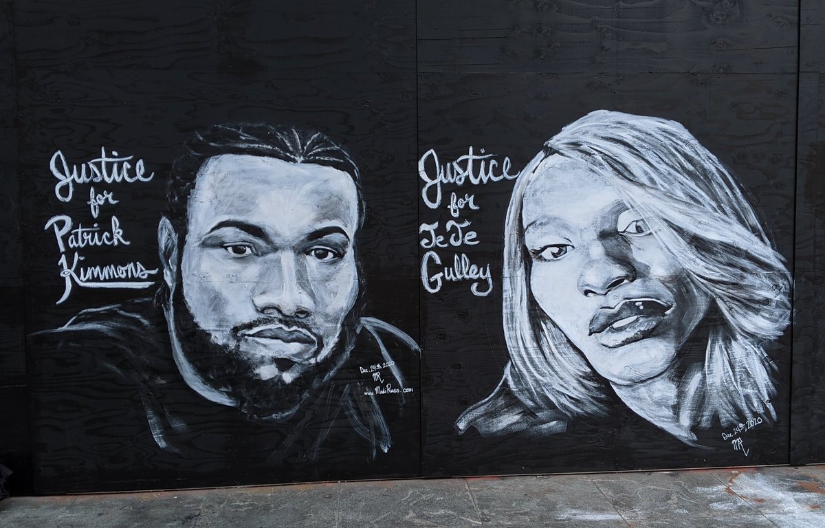 A landscape-oriented photo of a mural painted on a wall of plywood that's been painted black. There are two realistically rendered portraits painted in black, white, and grayscale. The left side portrait is Patrick Kimmons, a young black man with corn rows, beard and moustache, and long nose with flat nostrils. The right side portrait is TeTe Gulley, a black woman with light, straight, shoulder-length, layered hair, high cheekbones, a wide chin, and full, glossy lips. The ground below is paint stained concrete.