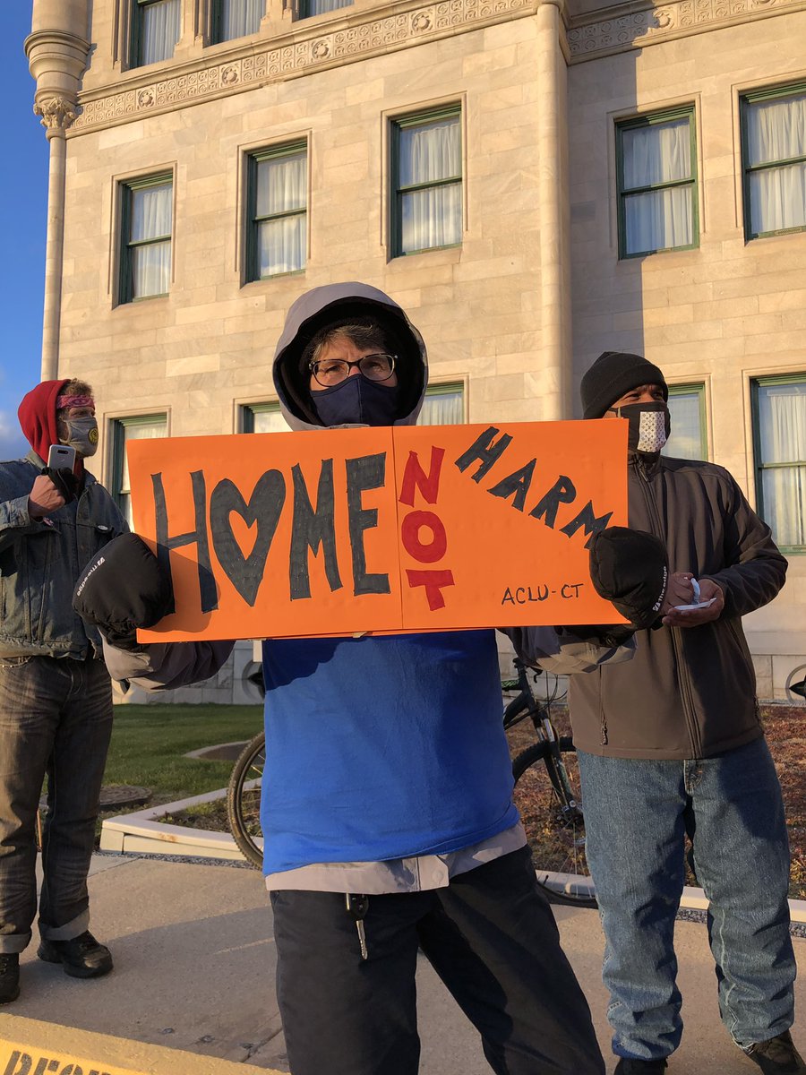 Person holding a sign that reads “home not harm”. Two other people in the background. One of them is holding up a phone.
