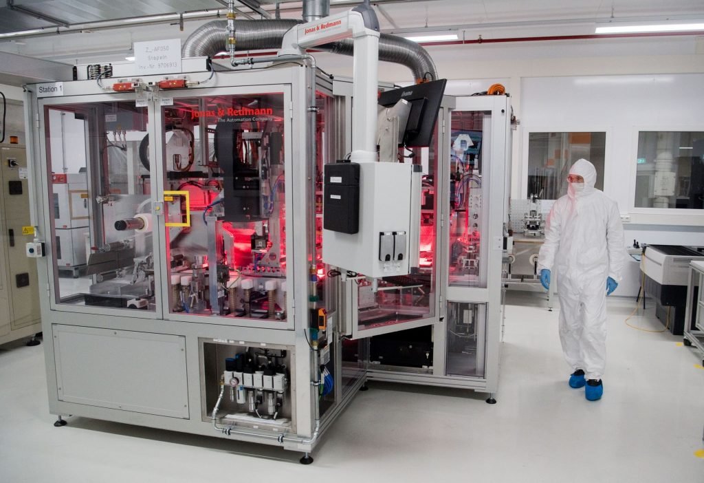 5/What is interesting is that: Cell production facilities are Class 7 clean rooms, while the worker doesn't wear the usual "bunny suit" protective gear. There's a barrier between the worker and the robot: likely separating clean-room air from unprotected air.