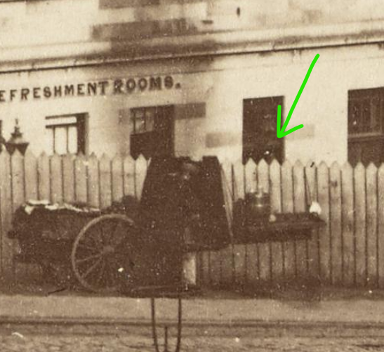 There are few people around, judging by the shadows it is the mid-afternoon so perhaps a Sunday? A single street seleer with a cart is present, they've set up a little booth, with a table hung from the picket fence with a brass-hooped barrel of some sort. Selling drinks?