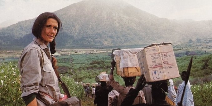 Photo of woman with long hair standing in front of a forest-covered mountain and fields. To the right, some people carrying parcels over their head.