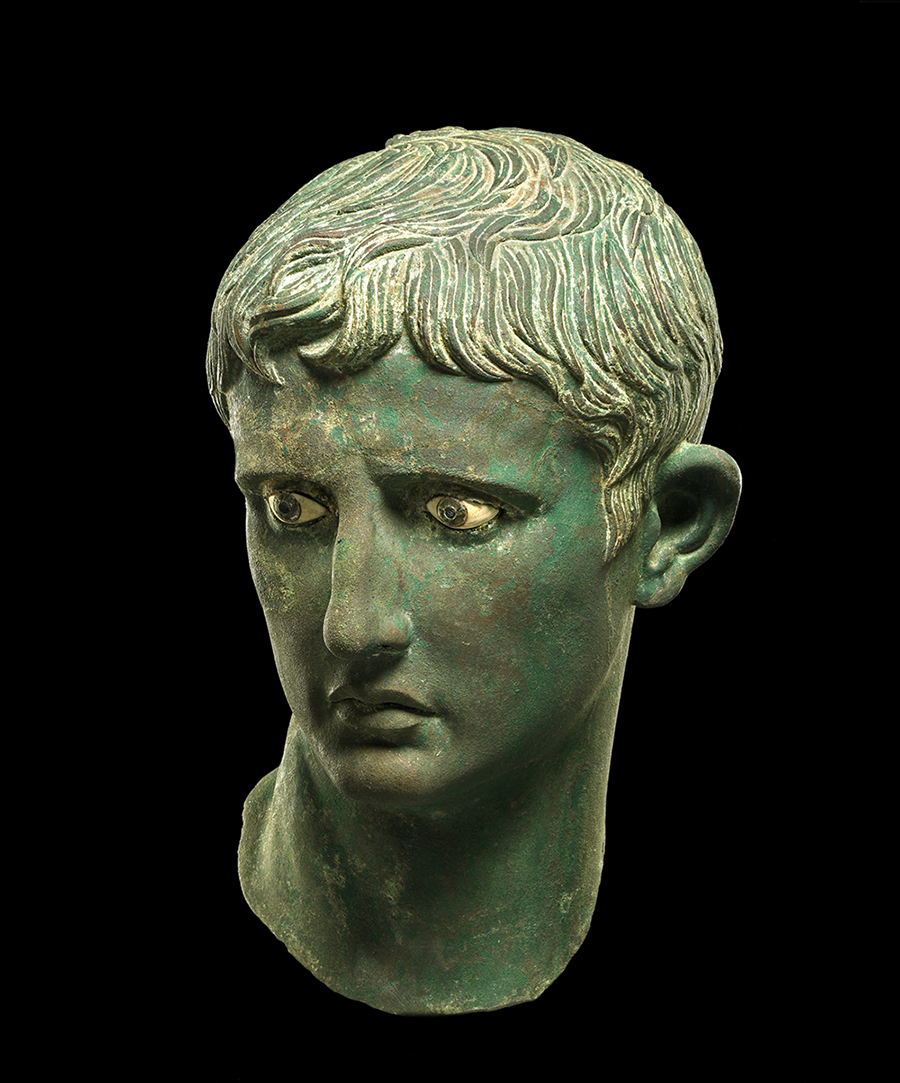 A greenish-grey statue of Augustus’ head against a black background. He has softly curled short hair, and inlaid white eyes.