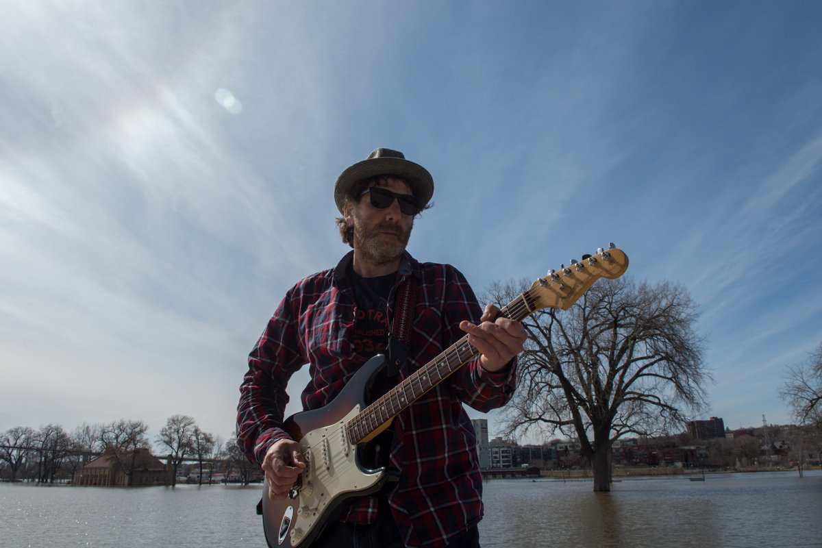 Dan Israel playing a Stratocaster outdoors, standing on the edge of a flooded Harriet Island part in St. Paul, Minnesota. Photo by Steven Cohen.