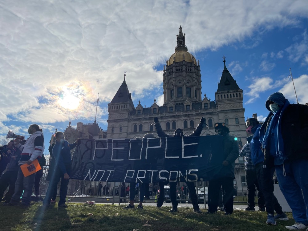 ACLU-CT smart justice leaders stand with a blue "people not prisons" banner in front of the CT Capitol Building. People are wearing masks and blue Smart Justice sweatshirts. One person in the center has his arms raised like Rocky Balboa, and one is holding a megaphone. In the background, the sun is shining behind the capitol dome.