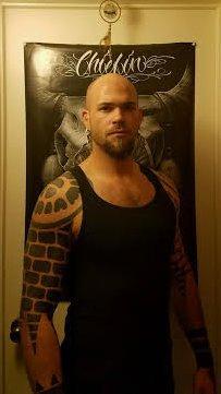 Photo of a bald man with a beard. He's wearing a black tank top and has tattoos covering his right arm.
