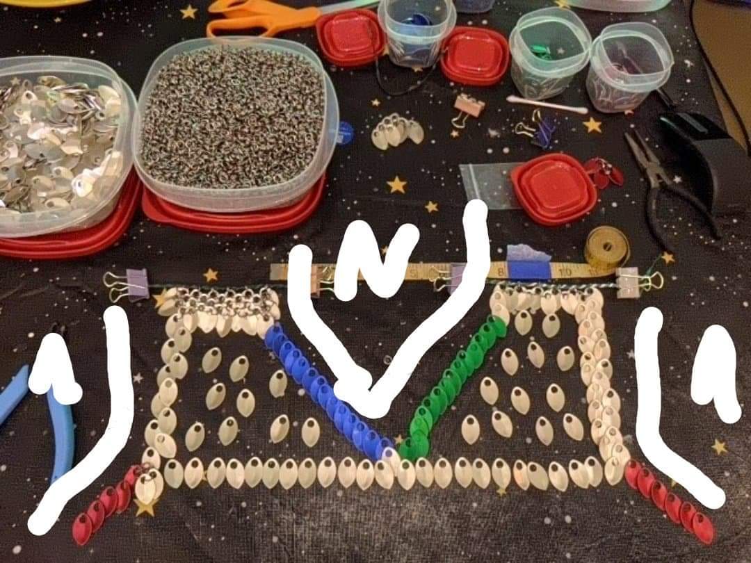 Showing the back side of scalemail armor some with rings, most just laid out to show how it will fit on the body. Red scales showing the pats that will go under the arms. Bins of color organized scales, rings line the table with other tools layed about.