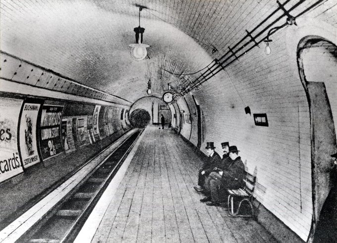 A black-and-white photo showing the platform of ‘British Museum Station’, with three men wearing top hats sitting on a bench, and one figure standing further down the platform.