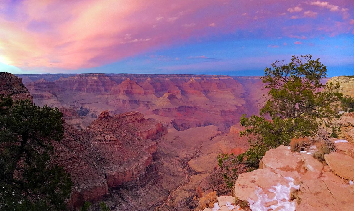 A vast canyon of colorful peaks and buttes taking on a pink hue from sunset light reflected from the clouds above. NPS/M.Quinn