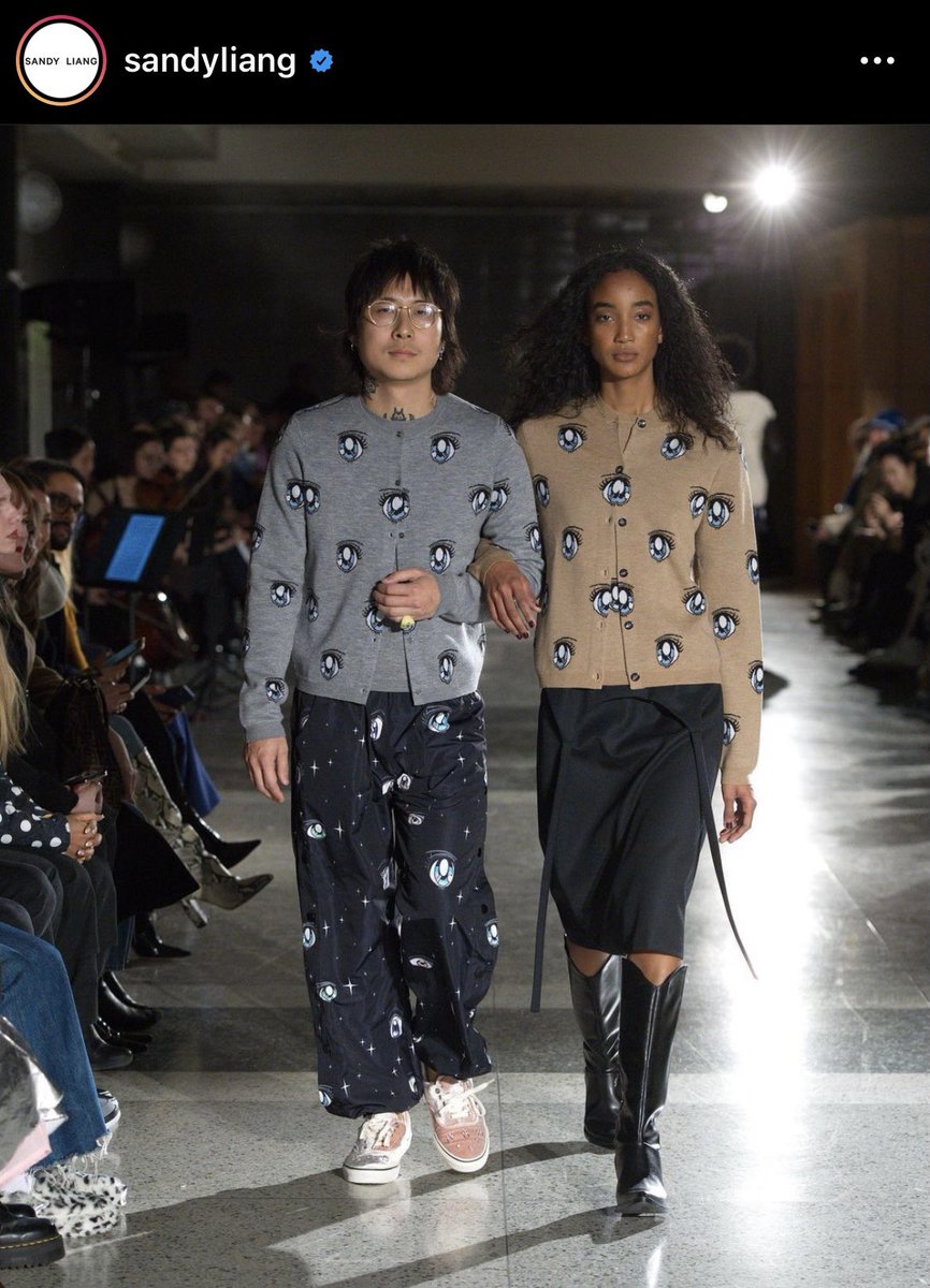 Two people walking down the runway, wearing matching cardigans patterned with cartoon eyes drawn anime-style