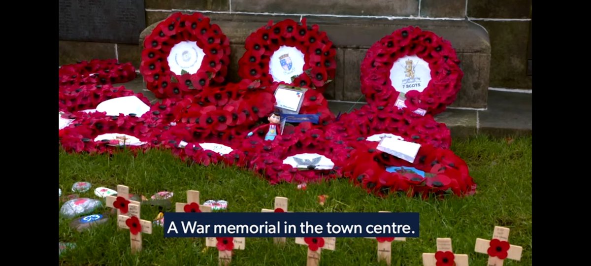 Wreaths of poppies at the foot of a war memorial. Small wooden crosses, each bearing a poppy, planted in the grass in the foreground.

Subtitles: "A war memorial in the town centre."