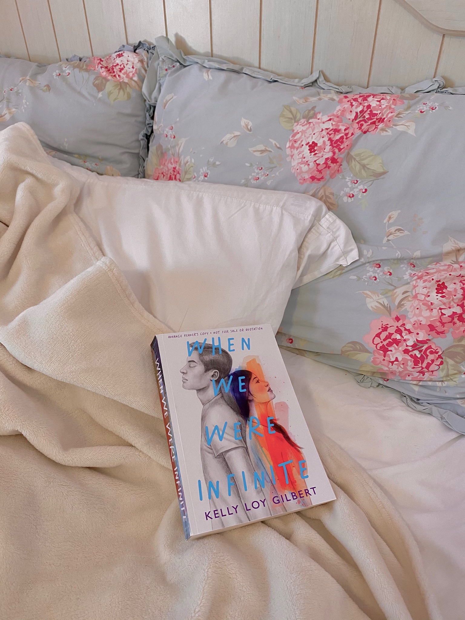 book When We Were Infinite by kelly loy gilbert sitting on top of off white blanket on and white pillow. behind are two blue pillows with pink flowers and a beige headboard
