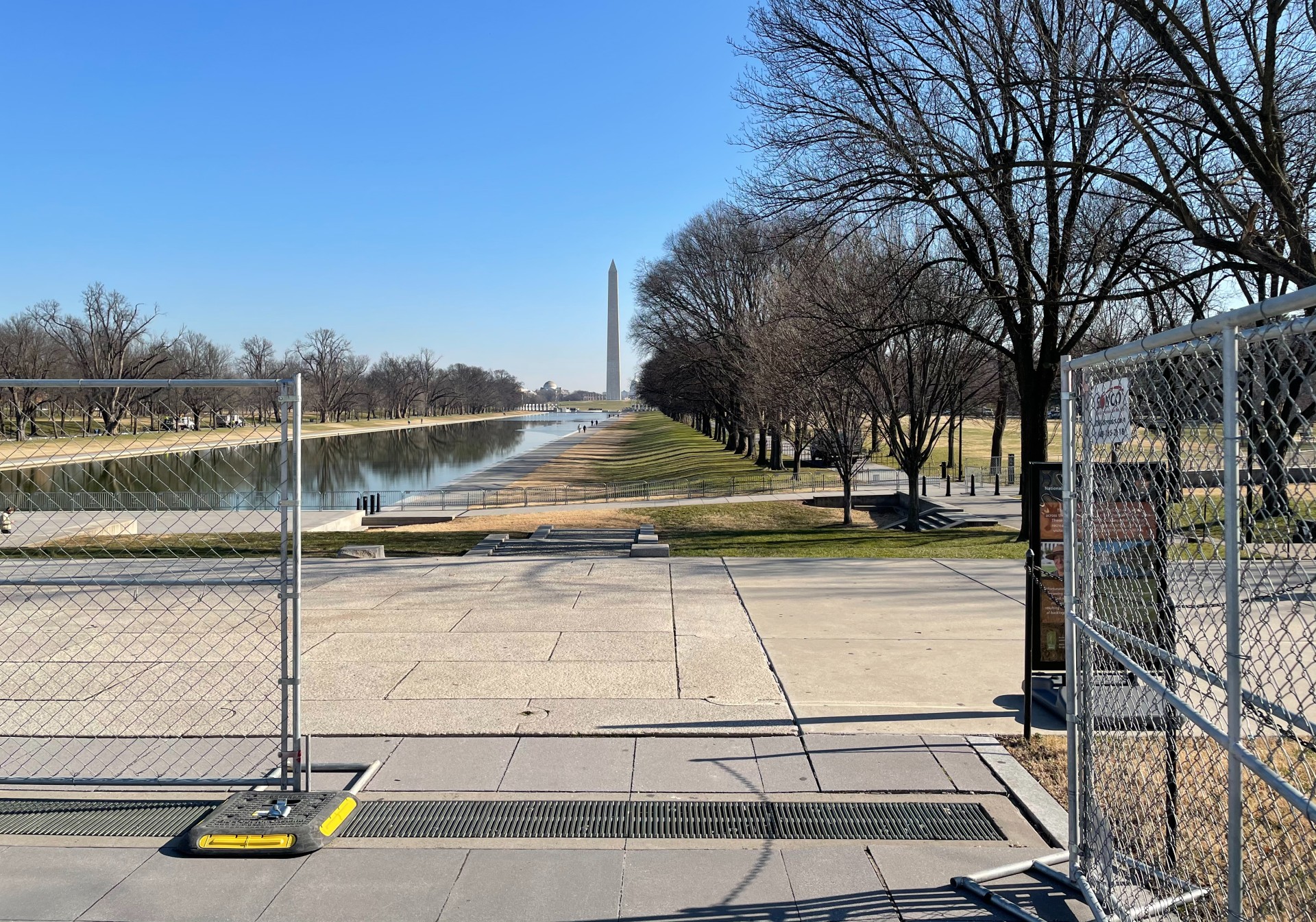 Chain link fence borders a walkway leading down to a long reflecting pool and the tower of the Washington Monument in the distance.