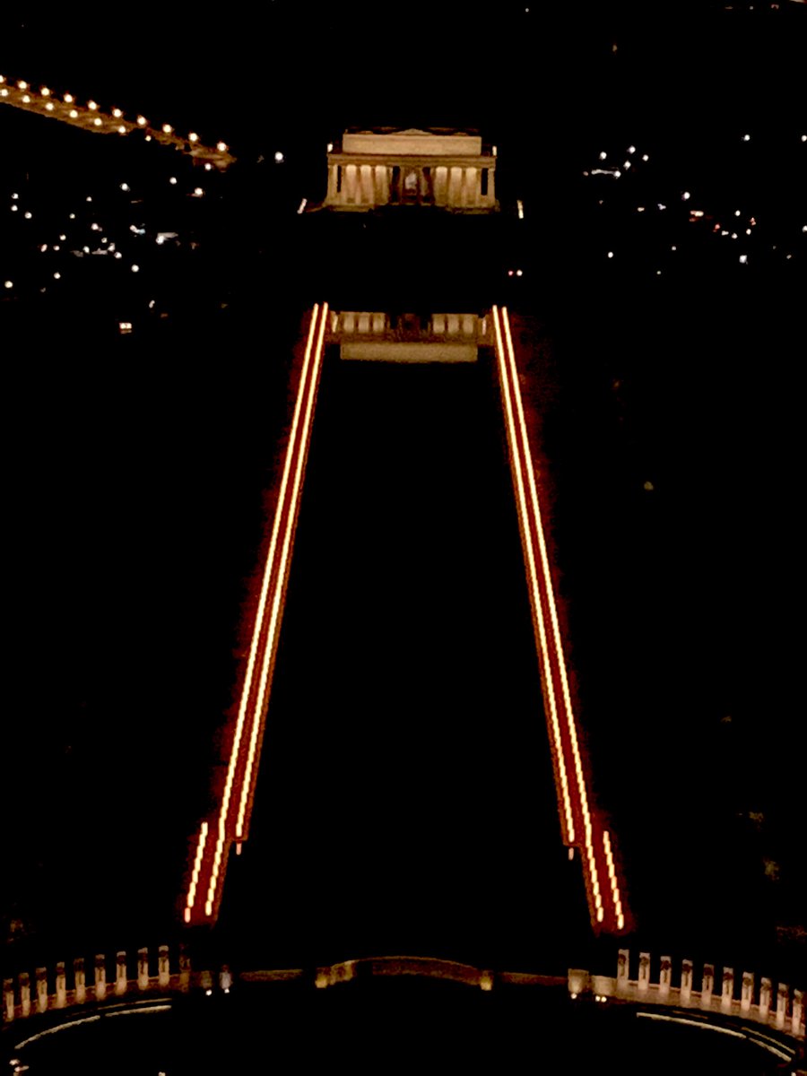 Two lines of light line both sides of a long reflecting pool with the flat roofed Lincoln Memorial at the far end on a dark night.
